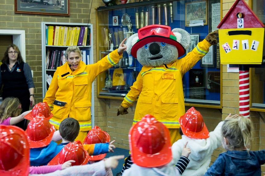 A woman and a person in a koala suit, both wearing firefighter uniforms, perform in front of a group of children.