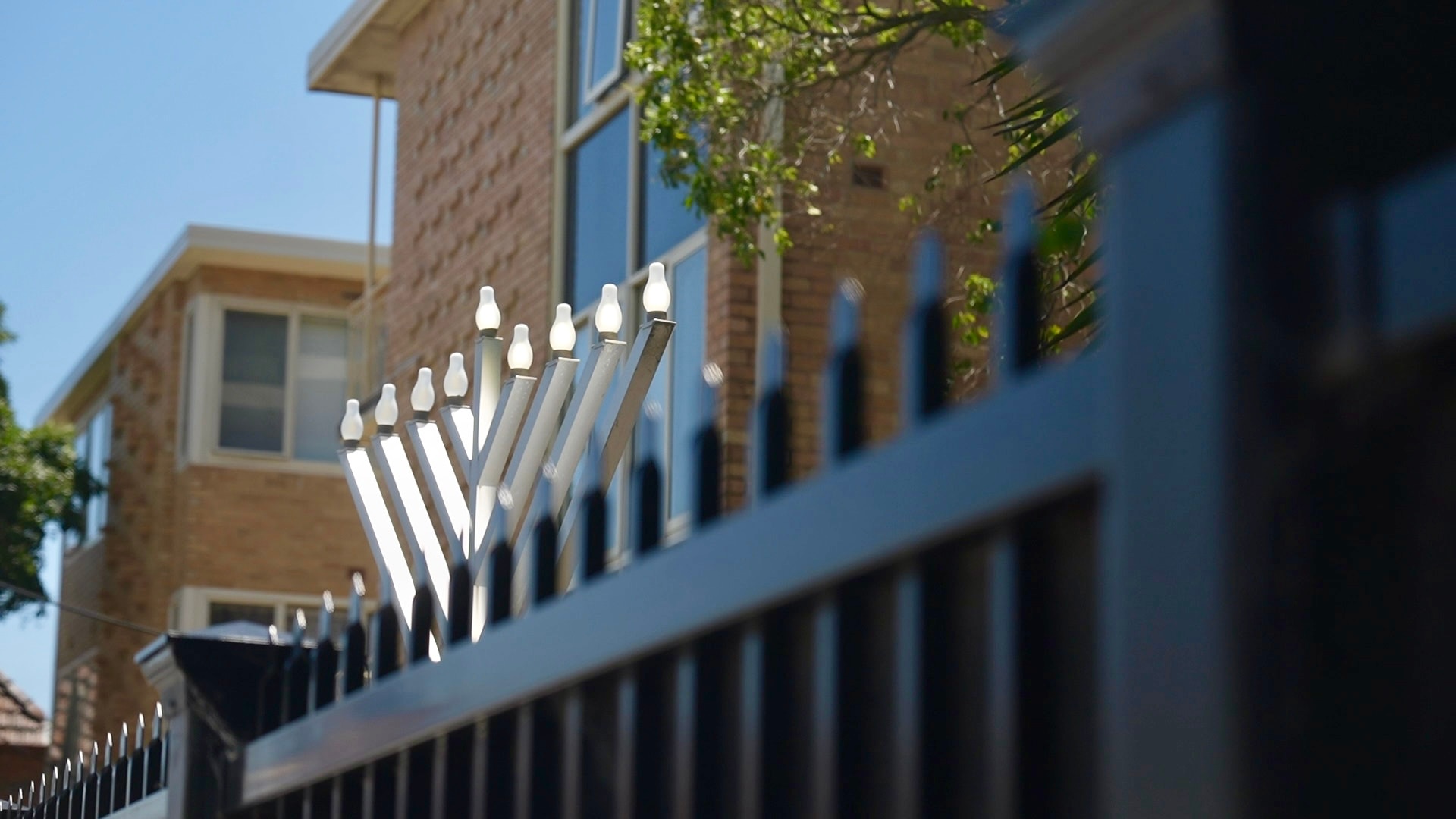 Uma alta menorá prateada com balões brancos pode ser vista no topo de uma cerca preta com pontas em frente a um prédio de tijolos laranja.