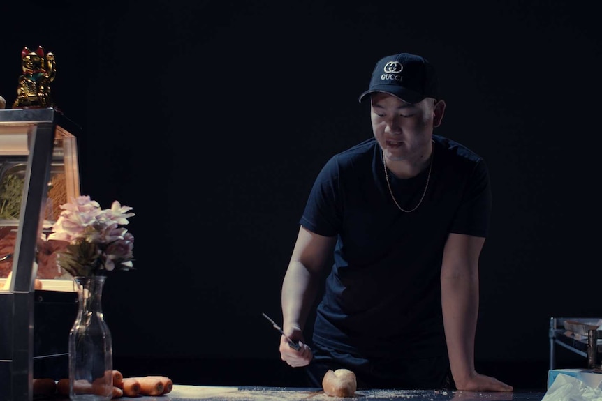 Young man in black pants, tee and cap stands at food prep bench holding knife, poised over a bread roll.