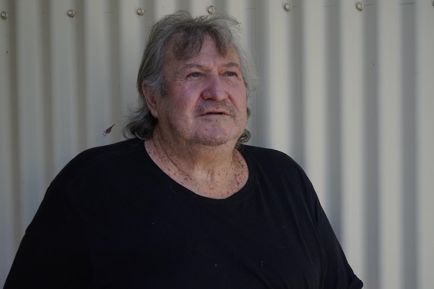 Robert Hargrave looks across the camera in front of a corrugated iron fence wearing a black t-shirt.