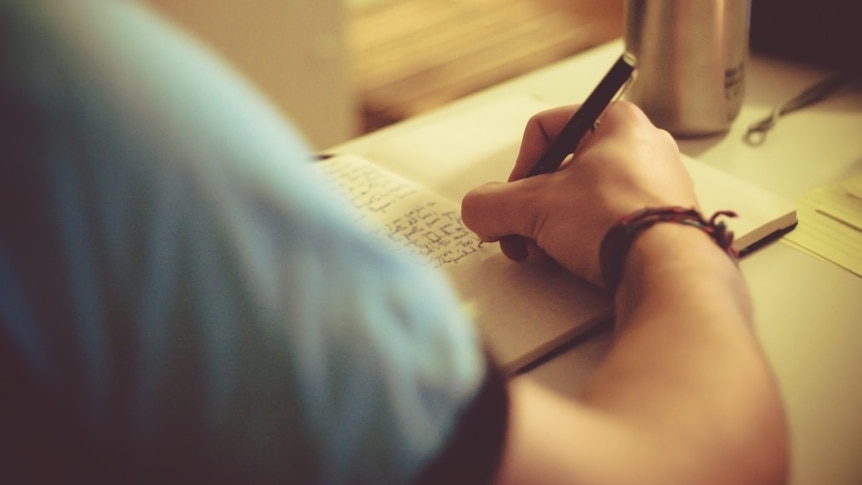 A photo taken over a man's shoulder shows him writing in a notebook.