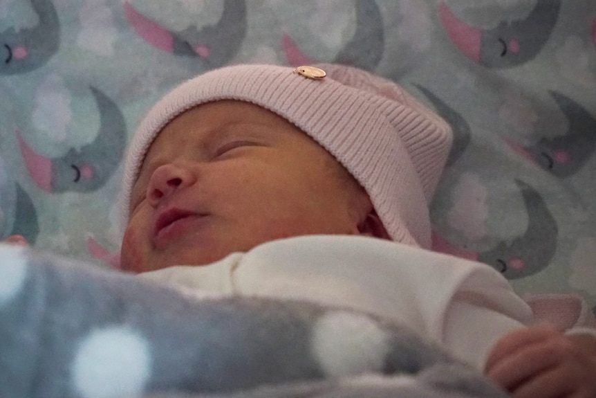 A small newborn wears a pink beanie in a hospital crib.