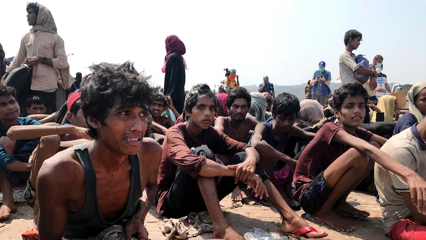 Dozens of malnourished ethnic Rohingyas sit on a beach. Closest to the camera is a man in a singlet crying.