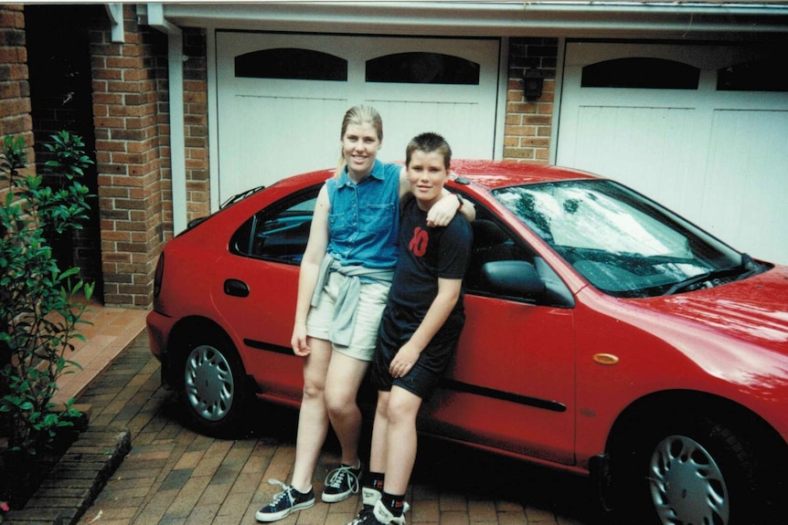 Wide shot of a young girl and boy leaning against a red car in a driveway.