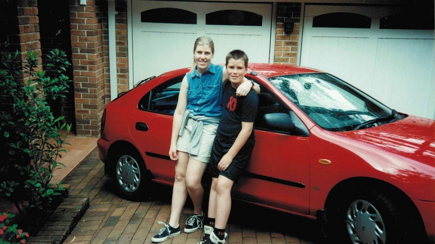 Wide shot of a young girl and boy leaning against a red car in a driveway.