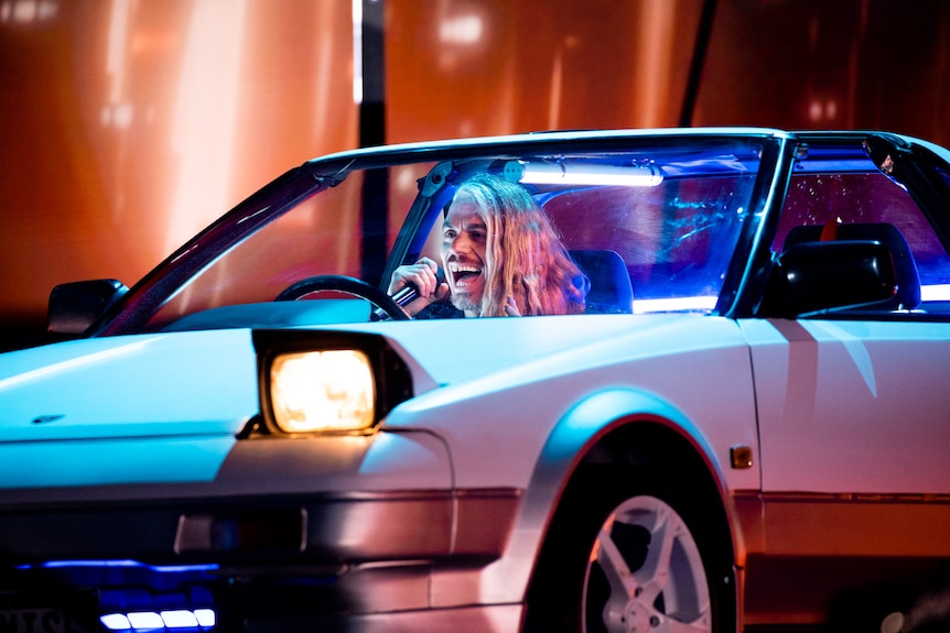 A man sings while sitting in a car with the headlights on, on stage during rehearsals for the Eurovision Song Contest.