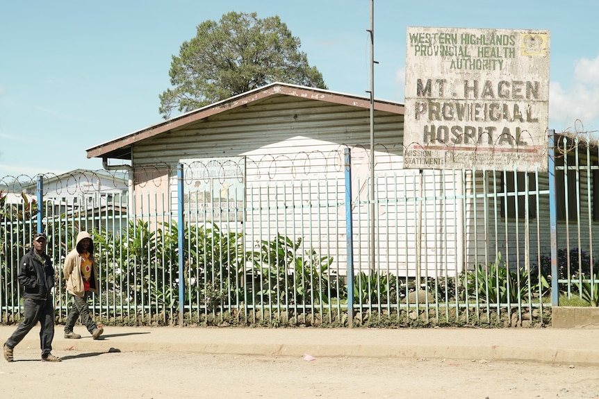 Two people wearing long pants and jackets walk by a sign that displays Mount Hagen Provincial Hospital.