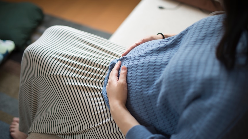 A pregnant woman pats her belly gently while sitting in living room.
