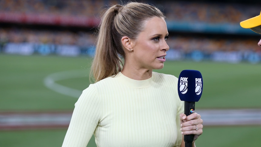 A TV presenter holds a microphone as she looks at a cricketer (at edge of shot) during a broadcast interview. 