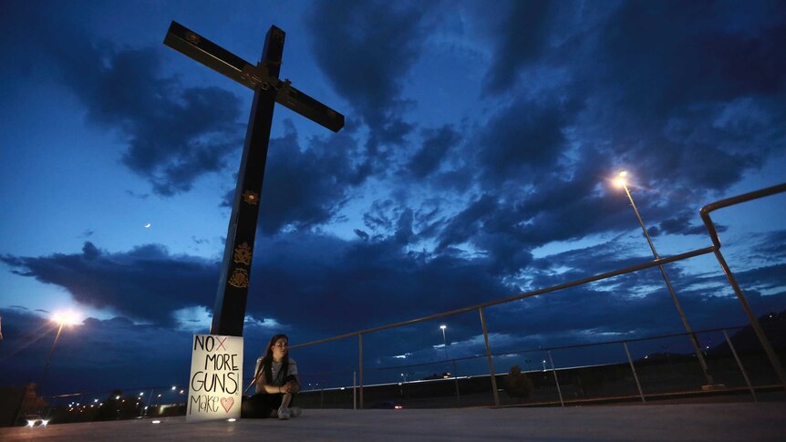 A cross against a dark blue sky with a woman sitting at its base with a sign saying No more guns, make love.