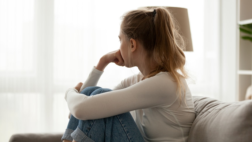 A girl with a pony tail, white top and jeans sits on a couch and looks out the window, she appears worried