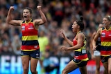 An AFLW player raises her arms in triumph as her teammates rush towards her.