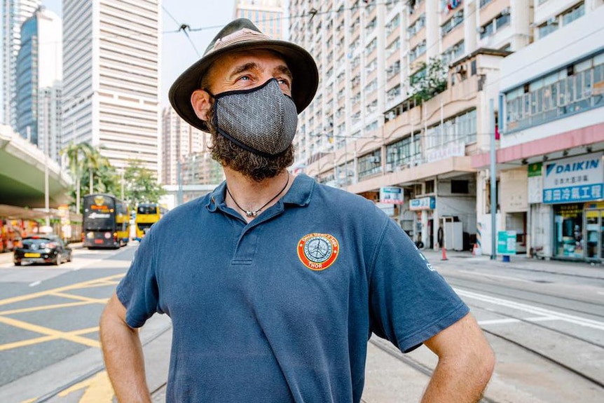 Thor wearing a face mask, a blue t-shirt, hat, poses with his hands on his hips on a Hong Kong road.