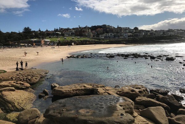 People swim in a calm ocean rock pool while others watch from the sand