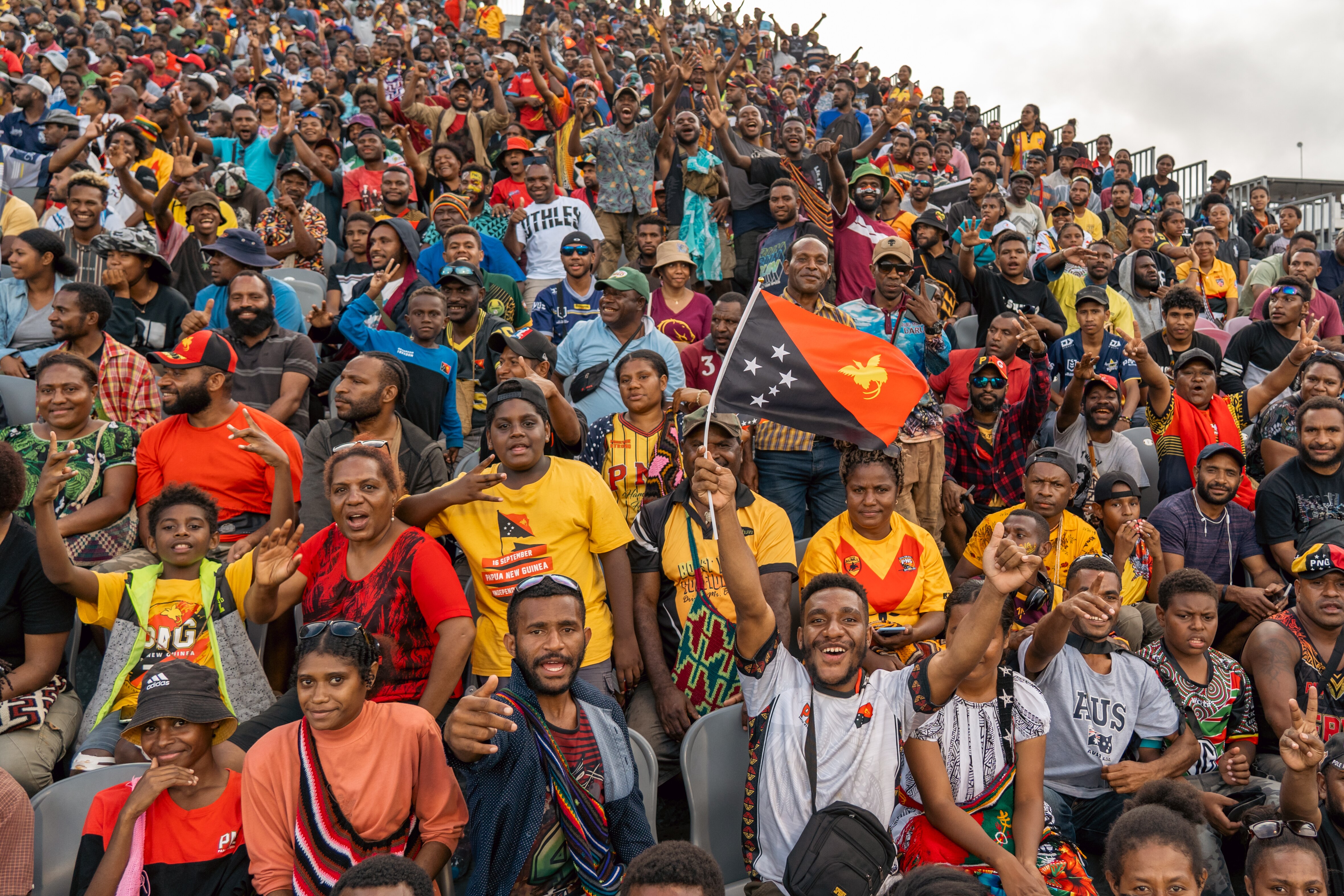 A crowd of NRL fans is pictured, they are cheering. Some hold flags and wear team jerseys.