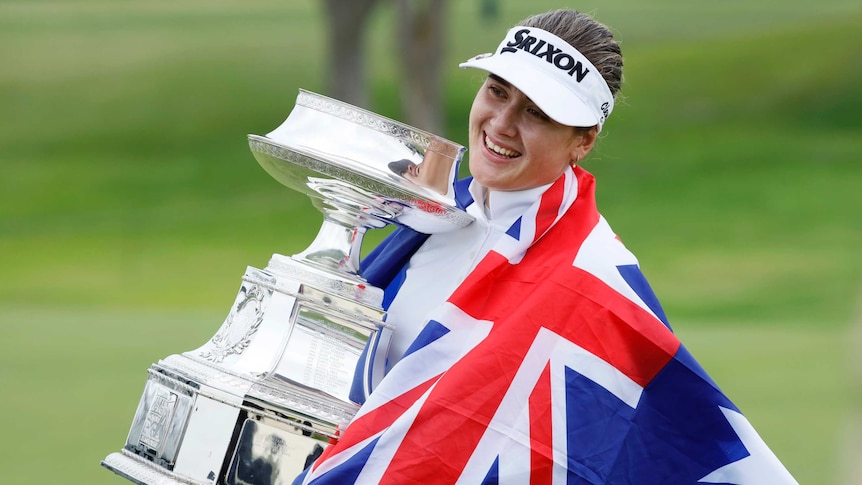 A golfer wrapped in an Australian flag holds the trophy after winning her first major title.