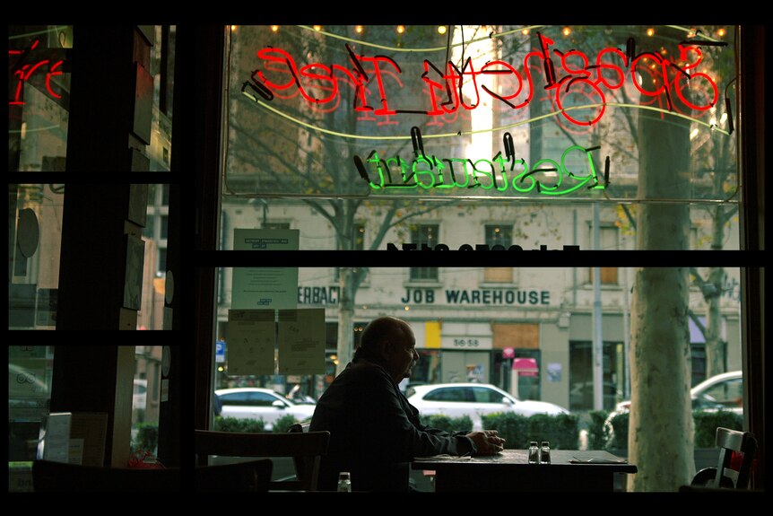 Danny, mostly in silhouette, sits by the window of a restaurant looking out at the street.