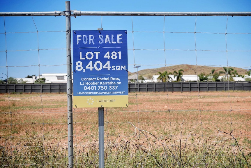 A small blue 'For Sale' sign on a wire fence in front of a vacant lot of land.