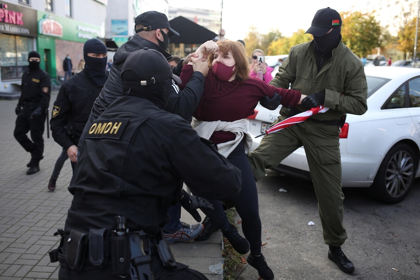 Three officers in hats and balaclavas grab a woman as they try to bring her to her knees