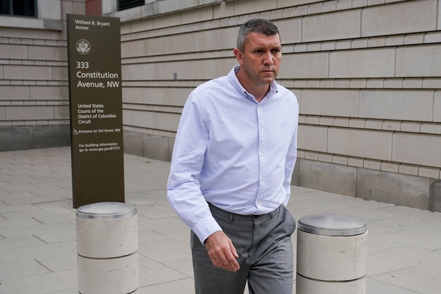 A man in a button up shirt looks past the camera as he walks outside a court building