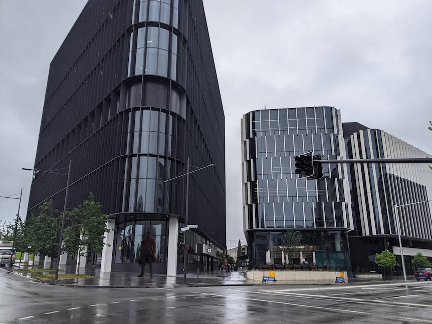 Two large new buildings rise up next to a street. Rain is falling.