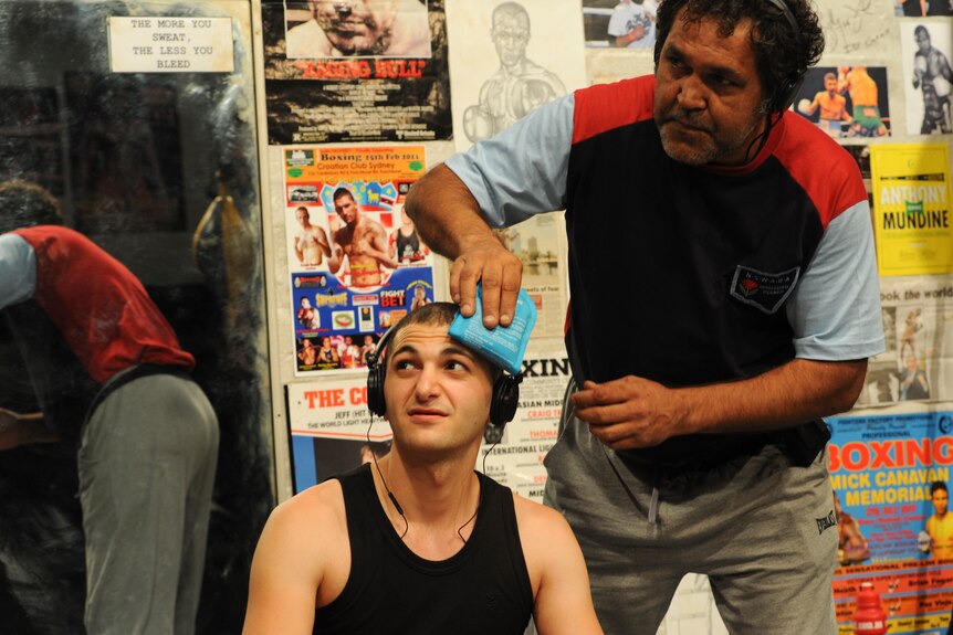 An older Aboriginal man places a wet cloth against the head of a seated Lebanese Australian man in his 20s