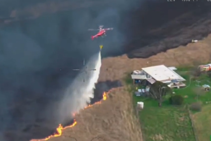 Aerial view of a red helicopter dropping water on a fire front that is approaching a home