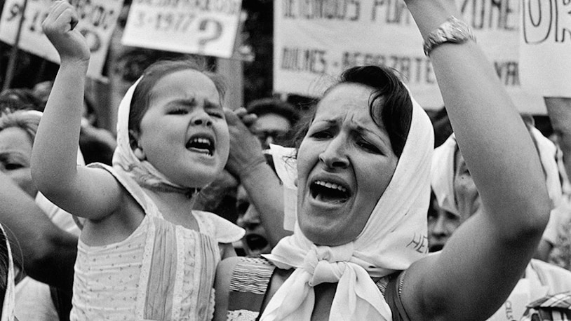 A black and white image shows a woman carrying a small child who shake their fists in the air in front of protest placards.