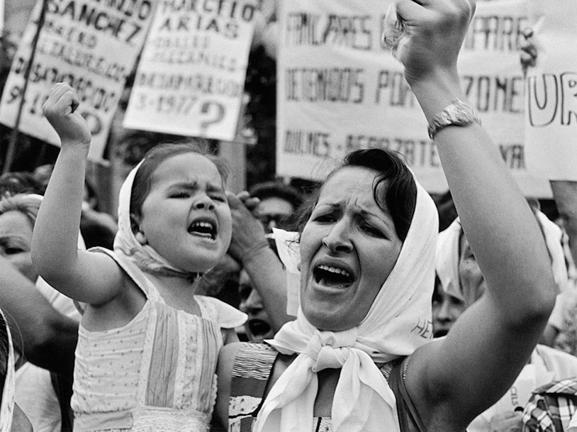 A black and white image shows a woman carrying a small child who shake their fists in the air in front of protest placards.