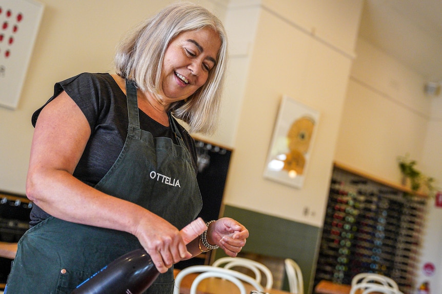 A woman wearing an apron opens a bottle of wine at a cafe table.