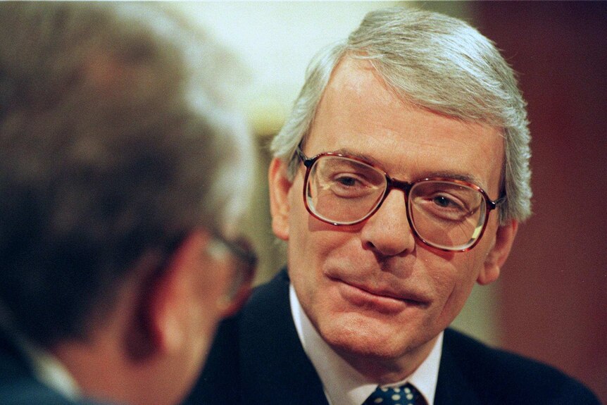 Sir John Major is shown close up behind the back of a blurred man's head with grey hair.