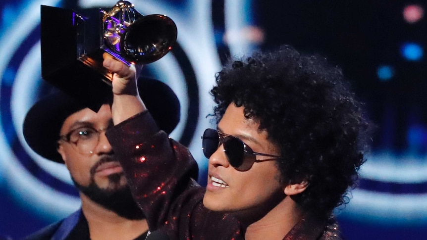 Pop star Bruno Mars holds up a Grammy award on stage and gives a speech from behind a microphone.