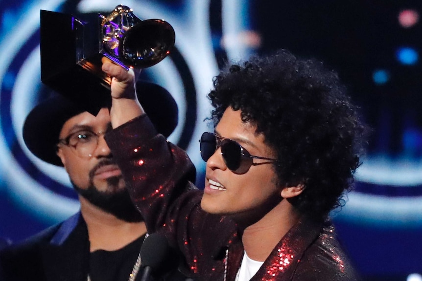 Pop star Bruno Mars holds up a Grammy award on stage and gives a speech from behind a microphone.