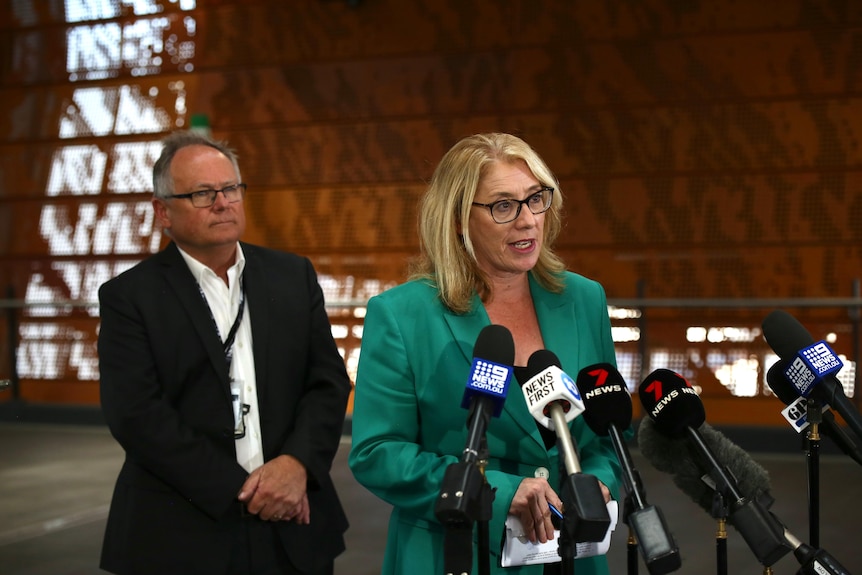 A woman in a green blazer speaks at a press conference while a man in a suit watches. 