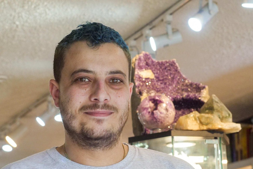 Jed Underwood has short, blue-dyed hair and is wearing a grey t-shirt. He is standing in front of a large purple crystal.