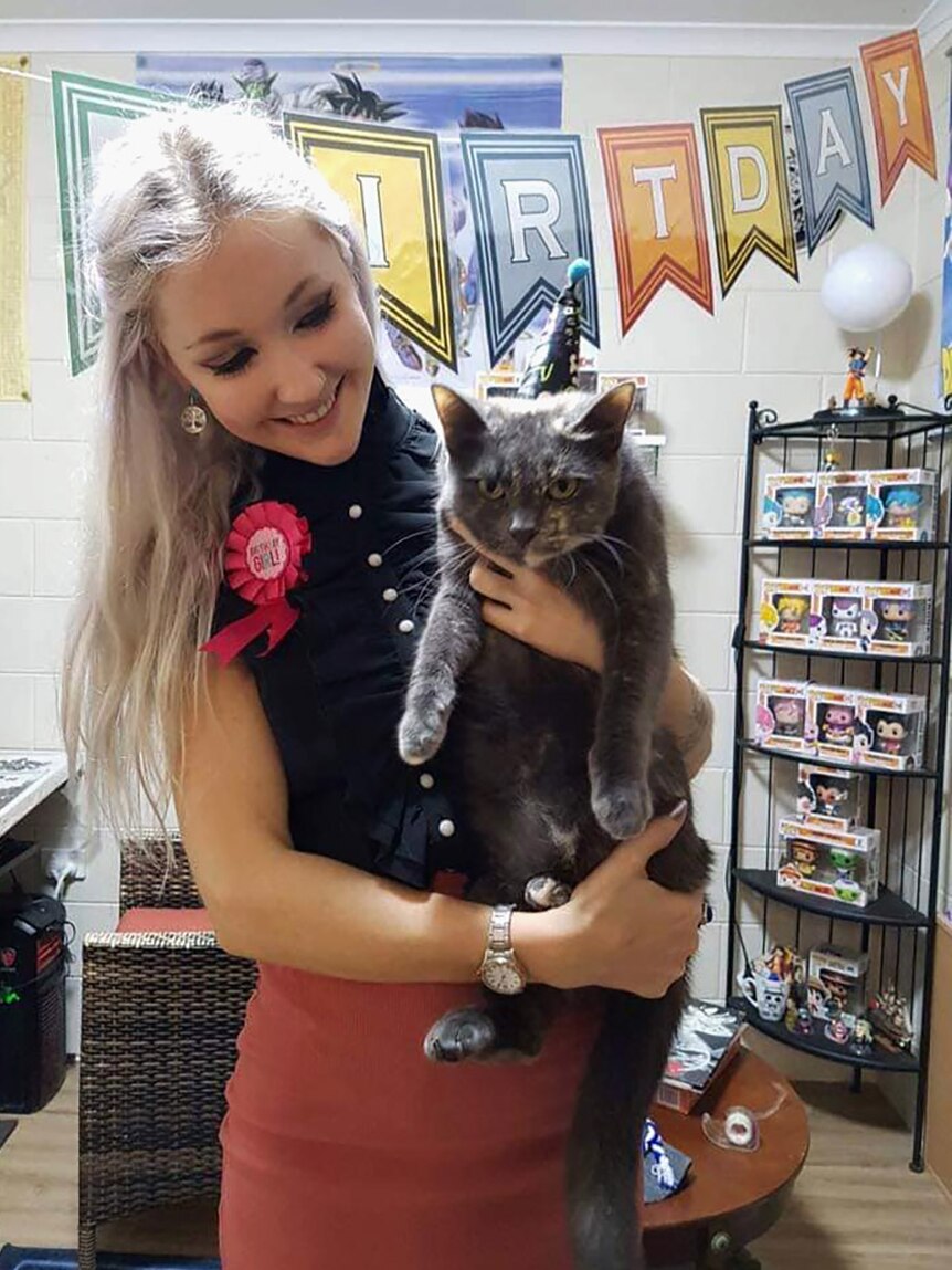 A smiling young woman holding a cat. A birthday banner hangs on the wall behind her.