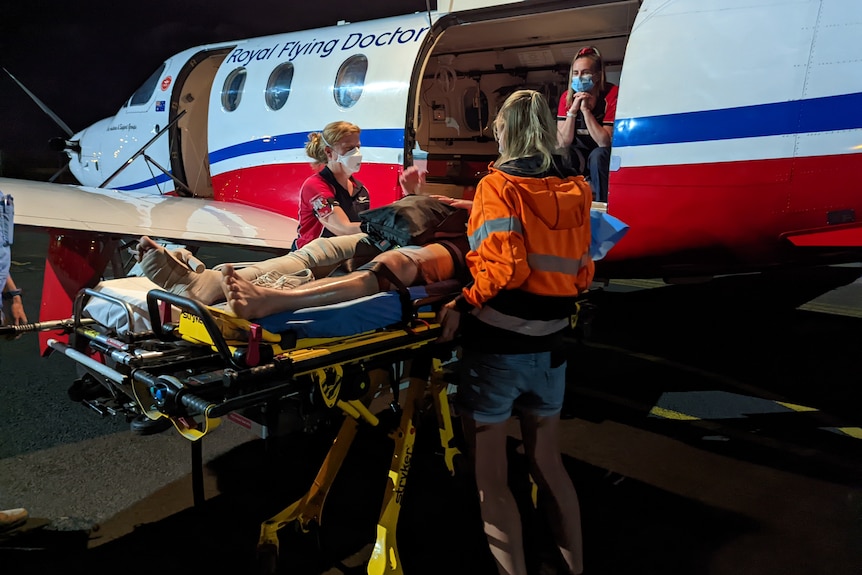 Two women wearing orange high-viz jacket, standing either side of medical stretcher next toRoyal Flying plane.
