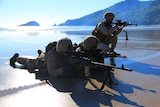 Military troops lie on a beach with guns pointed.