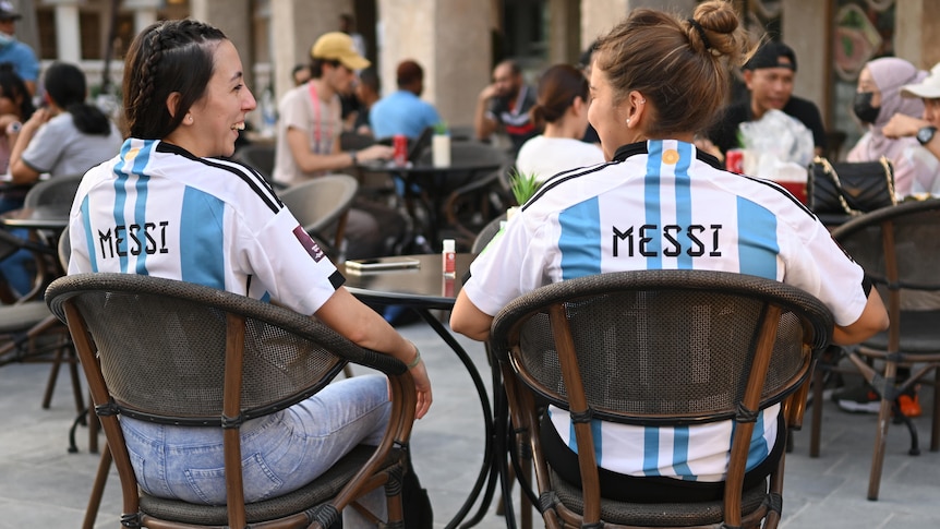 Two women wearing Argentina jerseys with "Messi" on the back sit at an outdoor table