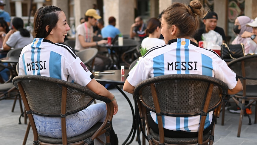 两名身穿阿根廷队球衣、背后有“Messi”字样的女子坐在户外桌子旁