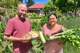 A couple smile at the camera holding vegetables and eggs with flowers and a cottage behind them