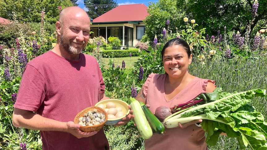 A couple smile at the camera holding vegetables and eggs with flowers and a cottage behind them