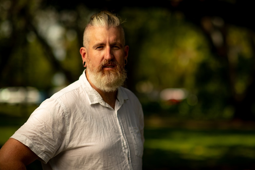 A man standing and looking at the camera, with greenery in the background.