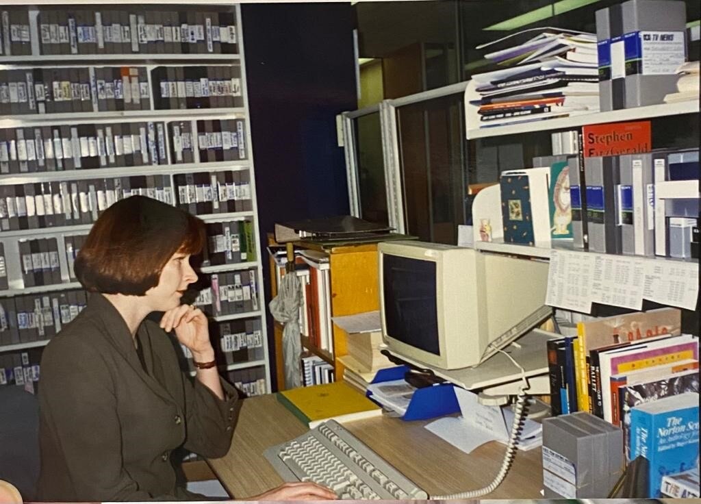 Woman sitting at old fashioned computer with tapes on a shelves behind her.