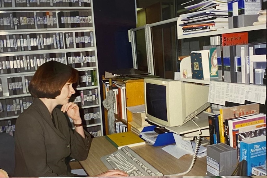Woman sitting at old fashioned computer with tapes on a shelves behind her.