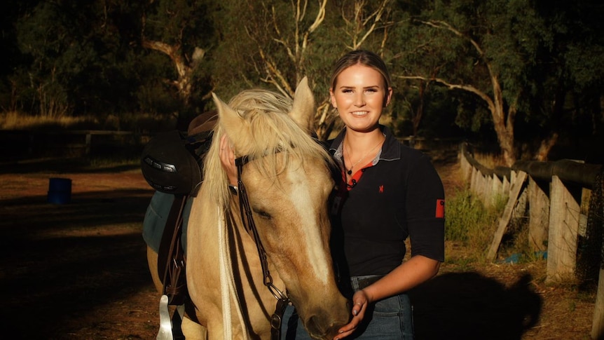 A blonde young woman pats a horse fondly.