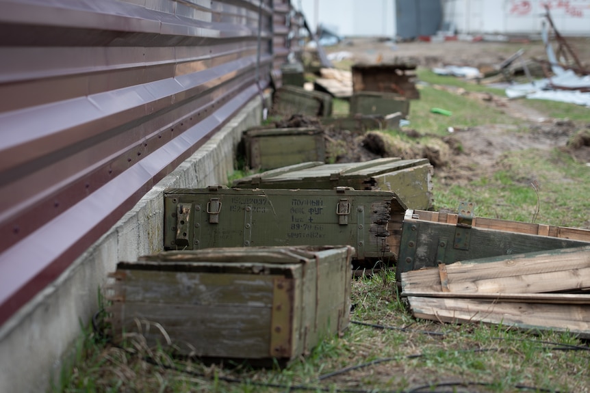 Discarded ammunition boxes lay on grass next to a fence.