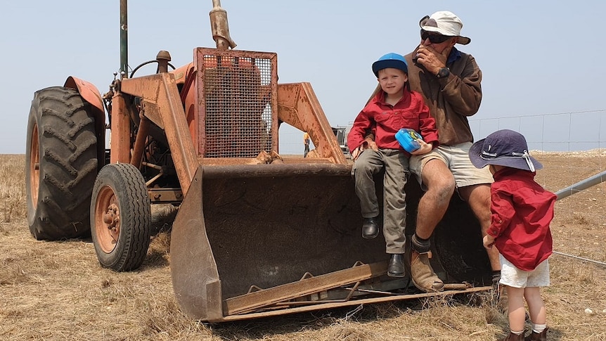 An older man sits on a burnt out tractor with a child in his lap, another child stands before him