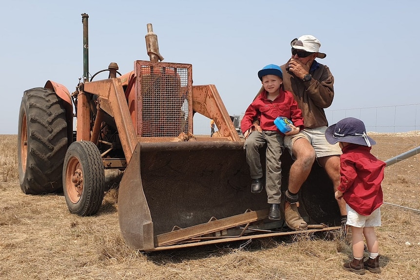 An older man sits on a burnt out tractor with a child in his lap, another child stands before him