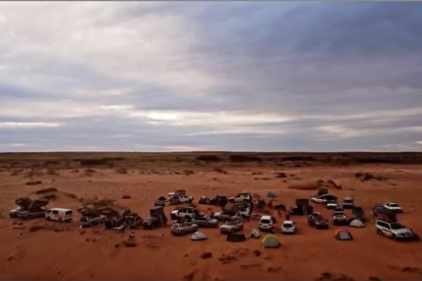 A long shot of about 30 cars on the beach under a clouded sky and red earth.